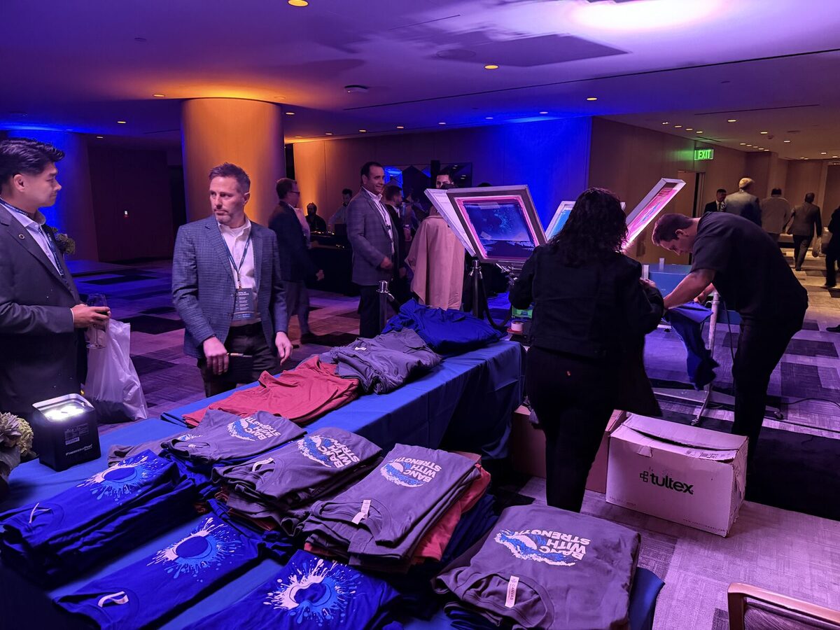 Folded custom-printed shirts in several colors stacked on a merch table while guests in business attire browse at a corporate reception