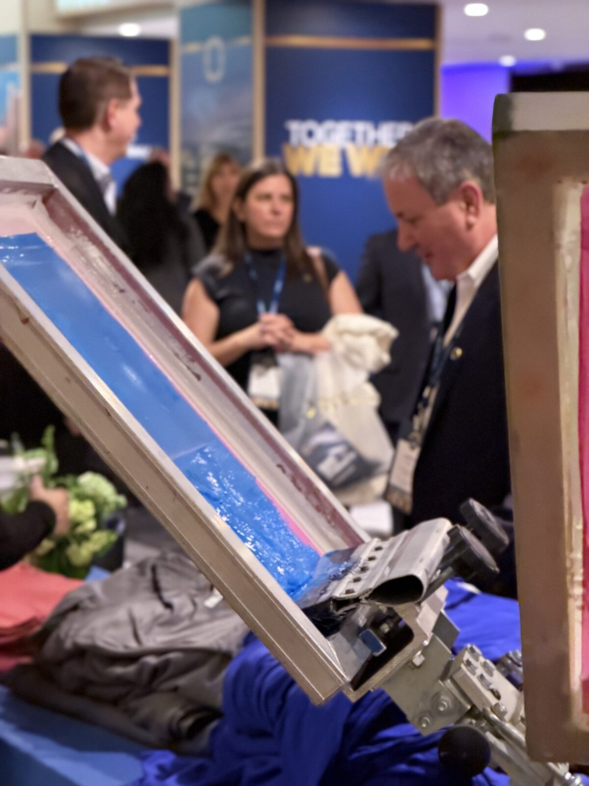 Close-up of a screen printing frame loaded with blue ink at an event station with conference guests in the background
